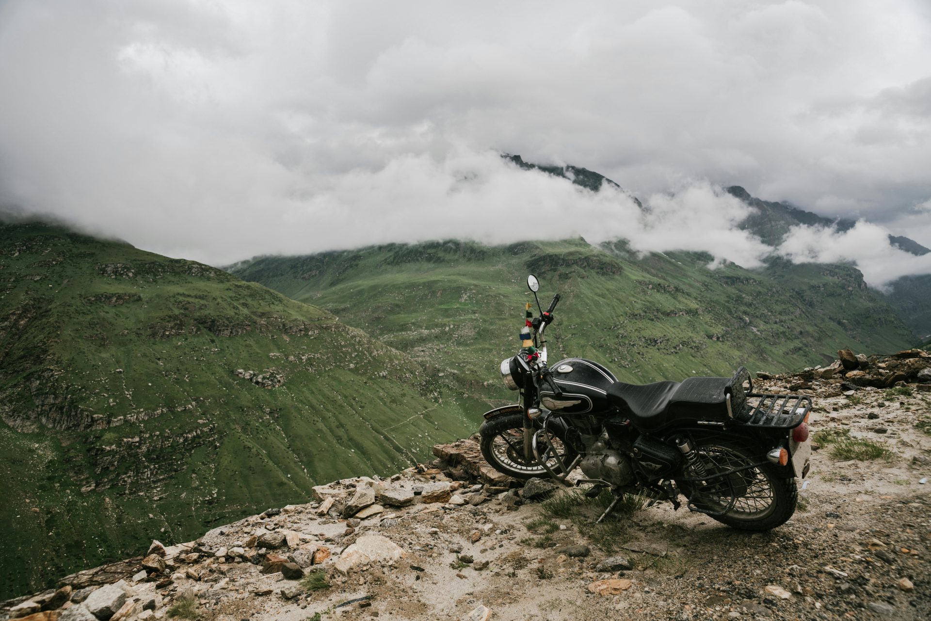A motorcycle parked on a rocky mountain cliff with a scenic view of lush green mountains and clouds.